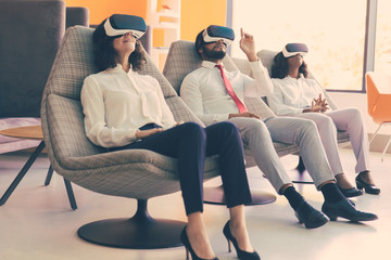 Obraz premium Team of excited business colleagues playing VR game. Man and women wearing virtual reality glasses, sitting and relaxing in motion chairs and touching air. VR studio concept