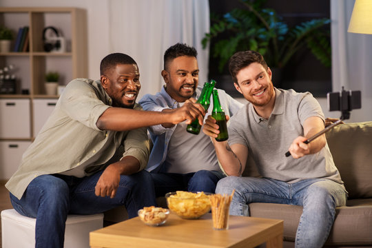 friendship, leisure and people concept - male friends taking picture with smartphone on selfie stick and drinking beer at home at night