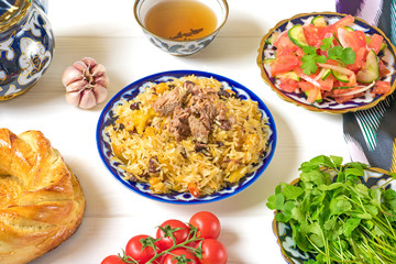 National Uzbek pilaf with meat, achichuk salad of tomato, cucumber, onion in plate with traditional pattern, cilantro, cherry tomatoes, garlic, bread tortilla - patir on white wooden table Top view