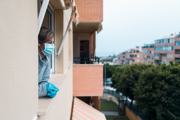 Woman confined at home by the government in prevention of the covid-19 coronavirus looking out the window at the deserted street in curfew.