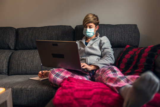 Concept: Woman in voluntary confinement with pajamas and surgical mask for prevention of coronavirus covid19 virus. She works at home using the sofa as a desk with a laptop and a smartphone.