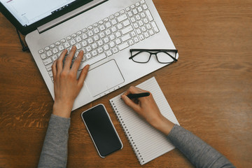 Work space with hands, laptop, notebook, glasses, pen, phone iPhone X. Top view on dark wood table background. Concept of mockup template.