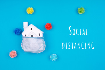 Cute little house covered with medicine mask and plastic balls aka viruses on blue background with Social Distancing wording. Epidemic, social isolation, coronavirus COVID-19 concept. Option with text