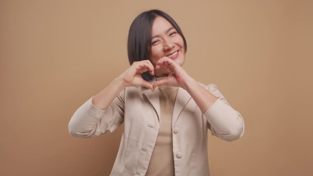 Happy asian business woman in love showing i love you sign language and looking at camera isolated over beige background. 4K video