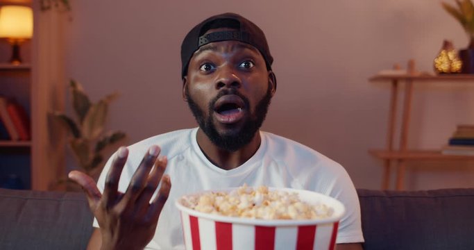 Handsome young guy making different face expressions while watching interesting film at home. Excited african eating popcorn from paper bowl while sitting on sofa in front of TV.