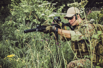 A military man or airsoft player in a camouflage suit sits in the grass and aims from an automatic rifle to the side