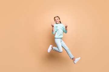 Fototapeta na wymiar Full length photo of pretty beautiful funny little lady jumping high up showing v-sign symbols rejoicing weekend wear blue sweater jeans footwear isolated beige color background