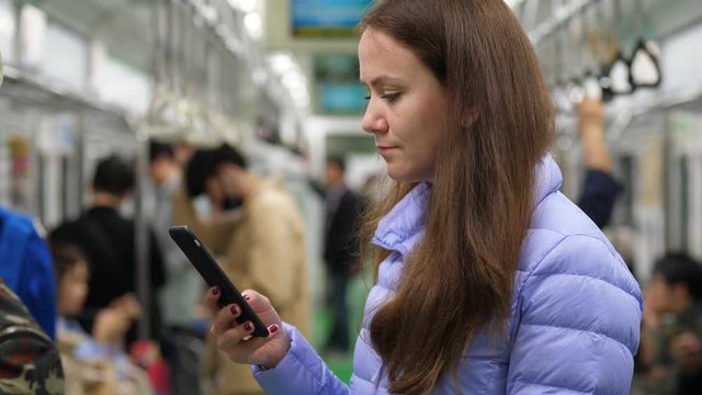 Adult woman standing at middle of subway car, texting by smartphone, blurred background. European tourist using metro of Seoul, rather empty train at evening time