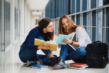 two pretty female students with books sitting on the floor in the university hallway