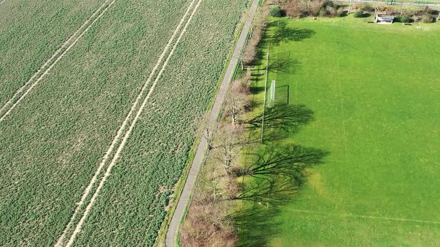 Aerial view of the flight over a cycle path between a field and empty football pitches to a road with a confluence and car traffic