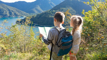 Fototapeta na wymiar Independent little children scouts with tourist backpacks stand on the shore of a mountain lake in the Alps and pave the way for a family boy scout weekend camping in the forest.