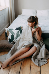 Beautiful happy sweet lonely girl woke up and wrapped in a blanket sitting on the bed in a spacious bright bedroom, resting and drinking coffee, holidays, weekends, hospital regime, cozy home