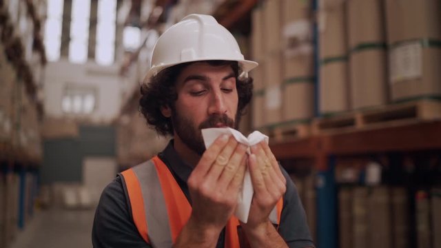 Sick allergic male factory worker in white hardhat and reflective jacket standing at warehouse blowing his running nose in tissue