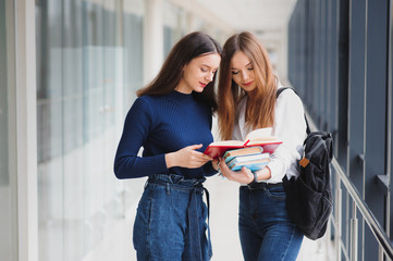 Two young female students standing with books and bags in the hallway University speaking each other.