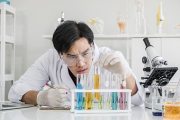 A male scientist with black hair wearing white coat and protective glassware looking serious holding up test tube in a laboratory setting with solutions and microscope.