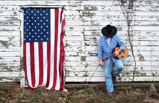 man looking down wearing cowboy hat leaning against old abondoned barn in the country holding acoustic guitar, American flag hanging on barn