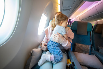 concept of flight with a child. little cute toddler jumping on her knees with a young beautiful mother in an airplane chair.