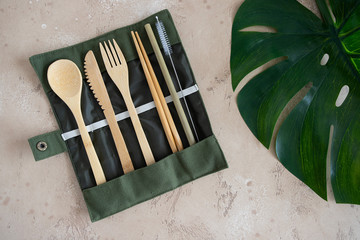 Eco friendly cutlery set, knife fork spoon, chopsticks  and bamboo straw with storage Bag on beige natural background with green monstera leaf . Zero Waste travel utensils