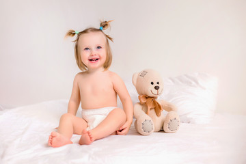 Toddler girl in diapers sits with toy bear on bed at home