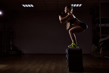 Fit young woman doing a step jump exercise. Muscular woman doing a box squat at the gym on dark backround