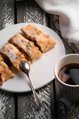apple strudel dessert on a white plate, cup of black coffee, old wooden backdrop