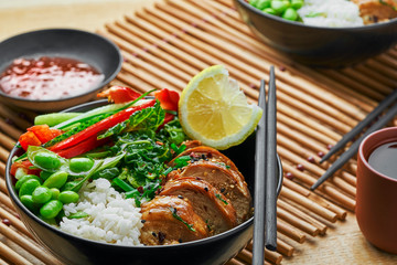 Teriyaki chicken fillet, fresh cucumber, red bell pepper, stew cabbage, edamame and basmati rice in black bowl. Two Asian Buddha bowls meal with green tea.