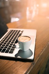 Side view of coffee cup placed on modern laptop. Wooden table retro
