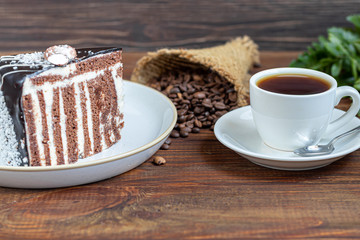 A piece of cake with chocolate icing. On a wooden background. In the background is a cup of coffee. Coffee beans are scattered all over the surface.