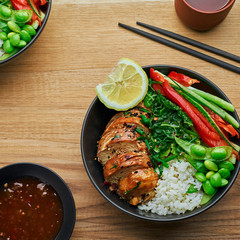 Teriyaki chicken fillet, fresh cucumber, red bell pepper, stew cabbage, edamame and basmati rice in black bowl. Two Asian Buddha bowls meal with green tea cups. Top view, directly above.