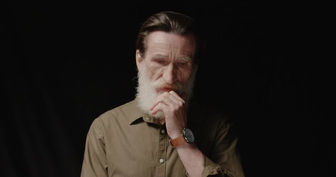 Portrait of handsome Caucasian old man on black background with serious face indoor. Thoughtful senior male model with grey beard posing to camera with conceived look and thinking. Emotions concept