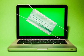 Laptop on a green background with a surgical mask on the monitor.  The computer shows a green background on the display.