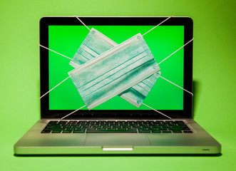 Laptop beech stands on a green background with surgical masks on the monitor display. Computer display shows a green background with surgical masks put on the monitor.