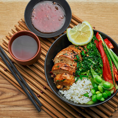 Delicious Buddha bowl with teriyaki chicken, fresh vegetables, edamame soy beans and basmati rice with green tea on a wooden table. Top view, directly above shot.