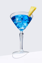 Blue Lagoon cocktail with ice cubes is contained in a martini glass with pineapple slice on the rim and isolated on the table edge. The showy illustrative picture is made on the gray background.