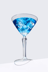 Blue Lagoon cocktail with ice cubes is contained in a martini glass and isolated on the table edge. The showy illustrative picture is made on the gray background.