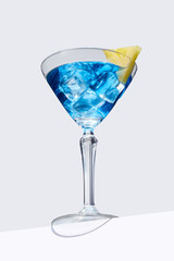 Blue Lagoon cocktail with ice cubes is contained in a martini glass with pineapple slice on the rim and isolated on the table edge. The showy illustrative picture is made on the gray background.