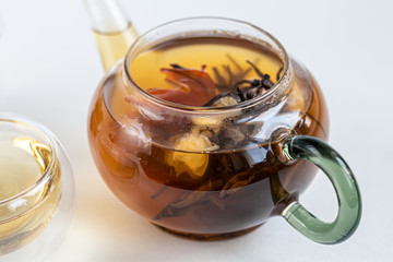 Blooming black tea in a glass teapot. On a light surface. Nearby are tea briquettes of leaves and flowers. In the background is a cake.