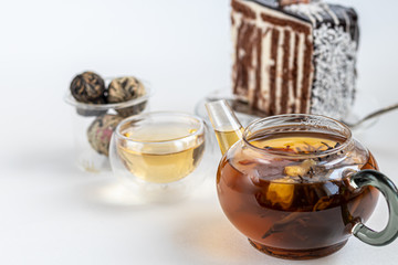 Blooming black tea in a glass teapot. On a light surface. Nearby are tea briquettes of leaves and flowers. In the background is a cake.