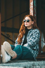 Stylish young beautiful girl in fashionable spring clothes with white sneakers sits on the asphalt