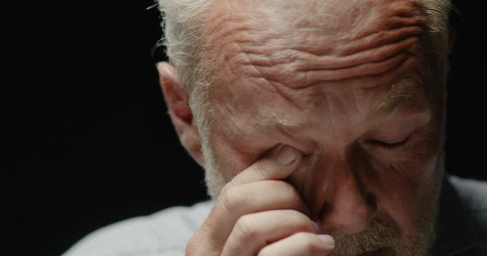 Close up portrait of sad handsome Caucasian old man with serious face indoor. Grey-haired bearded thoughtful senior pensioner on black background removing tear from eyes. Emotions concept