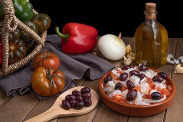 Esquexada in clay pot with ingredients and wooden spoon with some black olives.