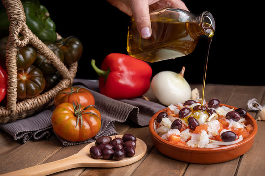 Esqueixada in clay casserole with ingredients and wooden spoon with some black olives watering with extra virgin olive oil.