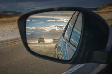 Cars seen in mirror traveling on misty road on transcanadian highway with rocky mountains in the background lit in the late afternoon sun. Epic travel or safety photo.