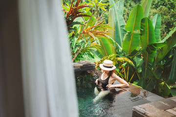 A young woman enjoys relaxing in a private pool at a luxury villa in Ubud, Bali, Indonesia. Girl resting in a pool on a background of green tropical plants. Luxury Holidays in Bali