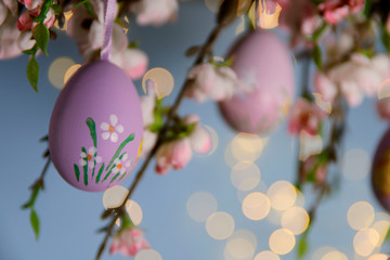Easter background. On a blue background with lights garland branches of a blossoming apple tree. Handmade silver easter eggs hanging on a branch. Close-up, horizontal, cropped picture, side view.