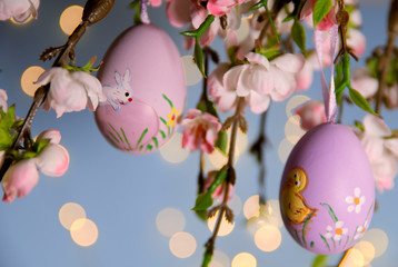 Easter background. On a blue background with lights garland branches of a blossoming apple tree. Handmade silver easter eggs hanging on a branch. Close-up, horizontal, cropped picture, side view.