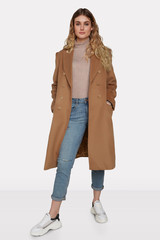 A blonde European lady with long wavy hair is posing on the white backdrop. The young woman is wearing white sneakers, blue jeans, beige jumper and a caramel-coloured unbuttoned coat.