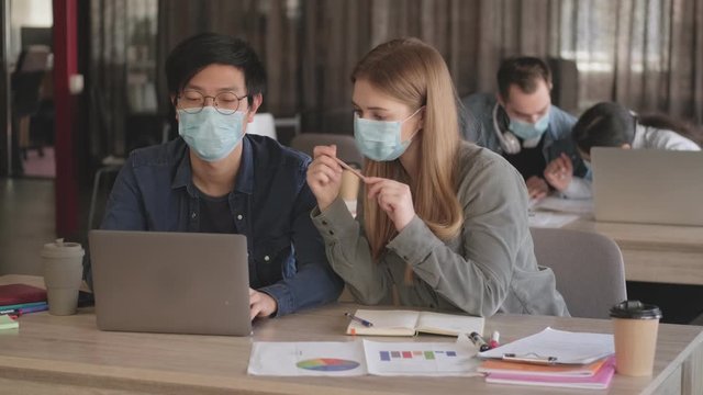 Young concentrated business people colleagues indoors in office talking with each other wearing masks because of coronavirus using laptop computer