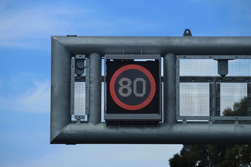 Overhead speed led sign on a bridge over a motorway (80 kilometres per hour) Electronic 80 in a red circle speed sign hanging over a freeway. Blue sky background.
