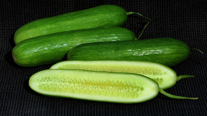 A group of green cucumbers one is sliced in half. The cucumbers are on a black rustic cloth background.